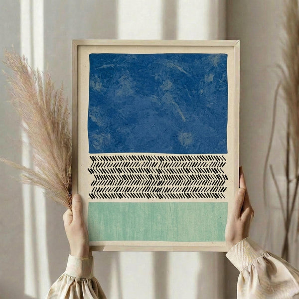 May include: Person holding a framed abstract art print with three horizontal sections - textured blue at top, black and white herringbone pattern in middle, and sage green at bottom. Light wood frame, neutral wall background, dried pampas grass visible to the left.