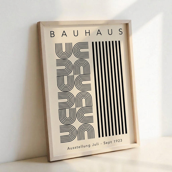 Framed vintage-style poster with cream background featuring black geometric typography reading "BAUHAUS" at top and "Ausstellung Juli - Sept 1923" at bottom. The design showcases bold striped letterforms arranged vertically alongside parallel black lines, displayed in a light wood frame leaning against a white wall.