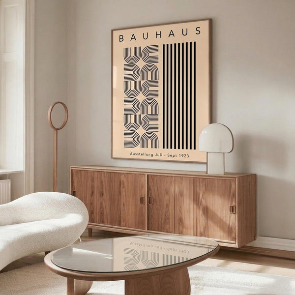 May include: Beige poster with black geometric text reading "BAUHAUS" and "Ausstellung Juli - Sept 1923" displayed in thin wooden frame above mid-century walnut credenza. Modern living room setting with cream upholstered chair, glass-top oval coffee table, white table lamp, and wooden floor lamp. Neutral walls with natural lighting.