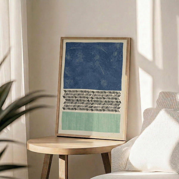 May include: Framed abstract art print with three horizontal sections - deep blue textured top section, black and white herringbone pattern middle band, and soft sage green bottom section. Print displayed on round wooden side table next to beige upholstered chair with white pillow. Natural lighting creates soft shadows across white wall, with partial view of plant leaves in corner.