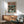 May include: Framed anime-style poster showing a figure in flowing cape facing a large mech robot in a rainy futuristic alley with Japanese text, displayed above mid-century modern walnut credenza with white ceramic lamp, next to teal upholstered armchair with wooden arms in bright living room with white walls and natural lighting.