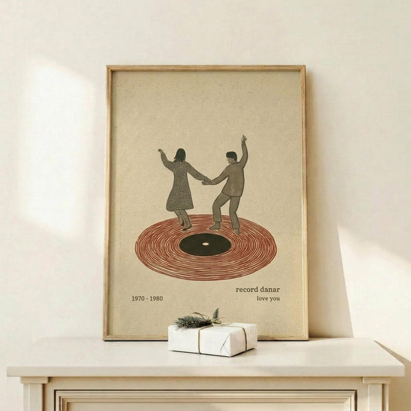 May include: Framed vintage-style poster on light wood shelf showing two silhouetted figures dancing on a red vinyl record, with "1970-1980" and "record danar love you" text below. Small white wrapped gift with evergreen sprig sits beside the natural wood frame against cream colored wall.