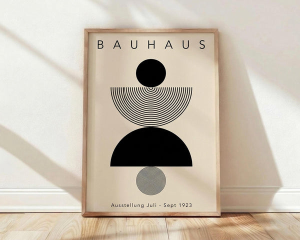 May include: Framed Bauhaus-style poster with geometric black shapes and concentric line patterns on cream background, displayed against white wall on light wood floor. Natural lighting creates soft shadows across the minimal composition featuring circles, semicircles, and radiating lines in modernist design.
