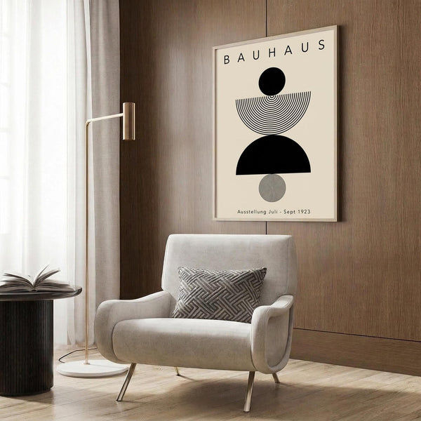 May include: Beige armchair with geometric patterned pillow positioned below framed Bauhaus poster on wood-paneled wall. Poster features black circles, semicircles, and striped patterns in minimalist design with "BAUHAUS" and "Ausstellung Juli - Sept 1923" text. Gold floor lamp and dark side table with open book complete the modern interior scene with neutral tones and clean lines.