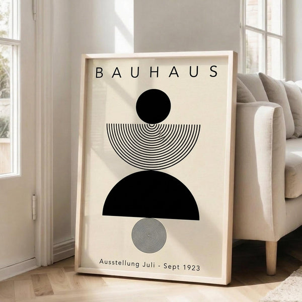 May include: Framed Bauhaus-style poster with geometric black shapes - solid circle, striped semicircle, solid semicircle, and small striped circle arranged vertically on cream background. Light wood frame leaning against white wall next to beige upholstered chair in bright, minimalist living room with large windows.