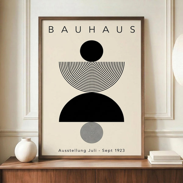 May include: Framed Bauhaus-style poster with geometric black and white design featuring circles and semicircles with concentric line patterns, displayed on wooden sideboard next to white ceramic vase and stacked books against cream paneled wall in modern interior setting.
