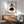 May include: Framed Bauhaus poster with geometric black and white shapes including circles and semicircles, displayed above a wooden sideboard with white ceramic lamp. Modern sage green upholstered chair with wooden arms sits nearby on wooden floor. Natural sunlight creates dramatic shadows across white wall in minimalist living room setting.