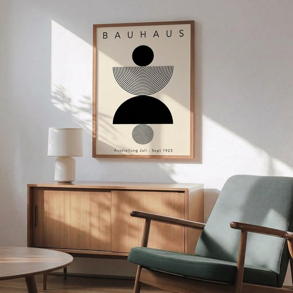 May include: Framed Bauhaus poster with geometric black and white shapes including circles and semicircles, displayed above a wooden sideboard with white ceramic lamp. Modern sage green upholstered chair with wooden arms sits nearby on wooden floor. Natural sunlight creates dramatic shadows across white wall in minimalist living room setting.