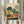 May include: Person in cream ruffled sleeves holding framed botanical print featuring various vegetables including leafy greens with orange roots, red tomatoes, cabbages, and radishes in vintage illustration style with green, orange and navy colors. Dried pampas grass visible beside figure against soft neutral curtained background.
