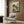 May include: Framed vegetable garden harvest illustration displayed above a beige armchair with geometric pillow in modern living room with wood paneled wall, gold floor lamp, and dark round side table with open book