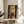 May include: Framed poster displaying four overlapping golden moon phases in vertical alignment against dark starry sky background, shown in thin gold frame on round wooden side table next to beige upholstered chair with white throw pillow, green plant leaves visible, soft natural lighting creating shadows on white wall