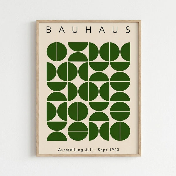 Framed poster with light wood frame featuring geometric green shapes arranged in a grid pattern on cream background, with "BAUHAUS" text at top and "Ausstellung Juli - Sept 1923" at bottom, displayed against white wall