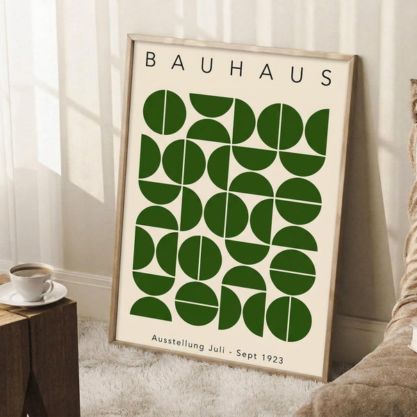 May include: Framed vintage-style Bauhaus poster with green geometric circles and semicircles arranged in a grid pattern on cream background, displayed in light wood frame leaning against white wainscoting wall next to wooden block with white teacup and saucer, on white textured rug beside beige knit throw blanket in bright natural lighting