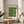 May include: Framed Bauhaus poster with green geometric circles and semicircles arranged in a grid pattern, displayed on a light wood round side table next to a beige checkered armchair. Soft natural lighting creates shadows on cream walls, with a plant visible in the corner. The vintage-style print features clean typography and minimalist design in forest green on cream background.