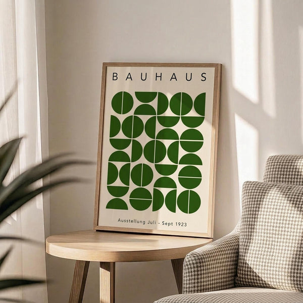 May include: Framed Bauhaus poster with green geometric circles and semicircles arranged in a grid pattern, displayed on a light wood round side table next to a beige checkered armchair. Soft natural lighting creates shadows on cream walls, with a plant visible in the corner. The vintage-style print features clean typography and minimalist design in forest green on cream background.