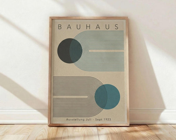 May include: Framed Bauhaus-style poster with geometric design featuring two circles in dark green and teal blue intersecting with curved parallel lines on beige background, displayed in light wood frame leaning against white wall on wooden floor with natural lighting