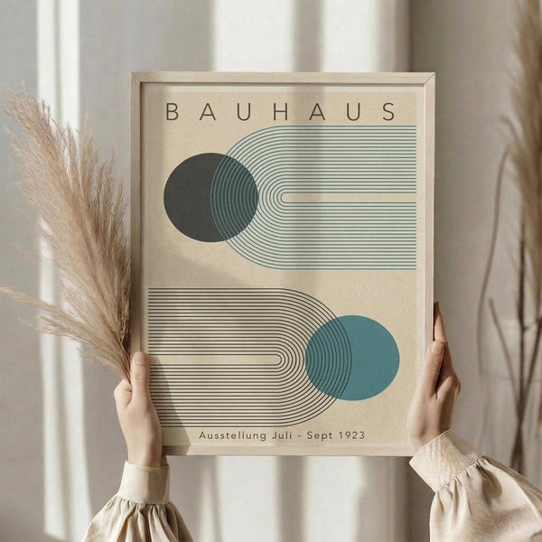 May include: Person in beige clothing holding framed Bauhaus poster with geometric design featuring two circles in dark green and teal blue connected by curved linear patterns on cream background, displayed next to dried pampas grass in soft natural lighting