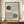 May include: Framed Bauhaus-style poster with geometric design featuring curved parallel lines forming flowing shapes around dark green and teal blue circles on cream background, displayed on wooden sideboard next to white ceramic vase and stacked books in modern interior with white paneled walls