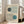 May include: Framed Bauhaus-style poster with geometric design featuring two curved linear patterns and circular elements in green and blue colors on cream background, leaning against white wall next to beige upholstered sofa in bright living room with large windows and natural sunlight streaming across wooden floors.