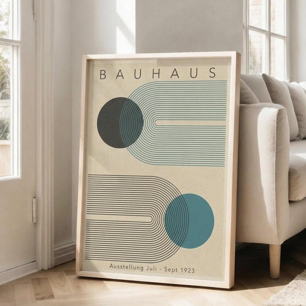 May include: Framed Bauhaus-style poster with geometric design featuring two curved linear patterns and circular elements in green and blue colors on cream background, leaning against white wall next to beige upholstered sofa in bright living room with large windows and natural sunlight streaming across wooden floors.