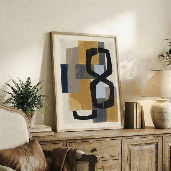 May include: Abstract geometric artwork in mustard yellow, navy blue, gray and cream tones displayed in light wood frame on rustic wooden sideboard. Modern art print features curved and angular shapes in contemporary style. Room scene shows neutral armchair, fern plant, ceramic vase, and stacked books creating warm, minimalist home decor setting with natural lighting.