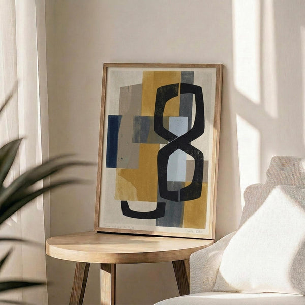 May include: Abstract geometric artwork in wooden frame displayed on round wooden side table next to beige upholstered chair. The print features overlapping rounded rectangular shapes in mustard yellow, navy blue, charcoal black, and light blue against cream background. Natural lighting creates soft shadows from nearby plant leaves across the minimalist interior scene.