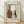 May include: Framed art print leaning against white wall on light wood floor. Print shows three pottery vessels - brown bottle with small dots, white vase with rust spots, and dark narrow vessel - surrounded by teal blue painted border on cream background. Natural wood frame with white matting. Soft natural lighting creates diagonal shadows on wall.