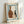 May include: Framed abstract art print featuring three pottery vessels in earth tones - a large brown bottle with golden dots, a white vase with terracotta speckles, and a dark narrow bottle. Blue painted border frames the still life composition on cream paper. Natural wood frame leans against white wall next to dried palm frond shadow.