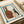 Abstract art print featuring silhouettes of two ceramic vessels - a brown pitcher with golden circular patterns and a white vase with rust-colored spots. The vessels are rendered in earthy tones against a cream background, surrounded by a hand-painted teal border frame. The artwork sits on white paper in an open art book or portfolio, with white ribbon visible at the edge, creating an elegant studio presentation.