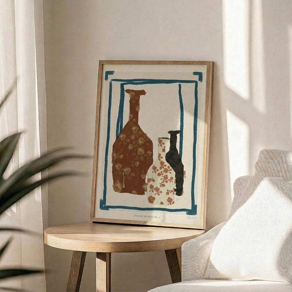 May include: Framed abstract art print featuring three stylized pottery vessels in rust brown, cream with terracotta spots, and dark teal, surrounded by hand-drawn blue border on cream background. Displayed on round wooden side table next to beige upholstered chair with white pillow, green plant leaves visible, soft natural lighting creating shadows on white wall.