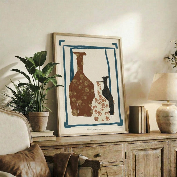 May include: Framed art print displaying three ceramic vessels in brown and cream tones within a blue painted border, positioned on rustic wooden sideboard alongside potted plants with large green leaves, stacked books, and ceramic vase in warm neutral living room setting with beige upholstered furniture.