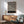 May include: Black and white swirling geometric pattern poster with Bauhaus exhibition text, displayed in thin wood frame above mid-century modern wooden credenza. Sage green upholstered armchair with wooden arms sits nearby, white ceramic table lamp on credenza. Sunlight casts diagonal shadows across white wall in minimalist living room setting.