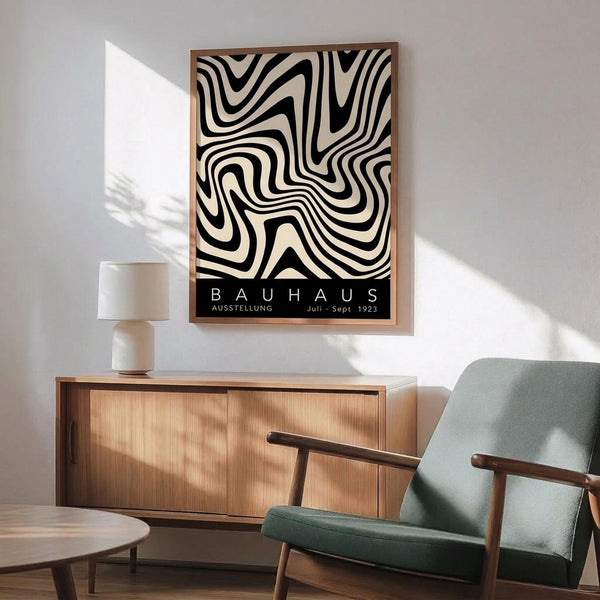 May include: Black and white swirling geometric pattern poster with Bauhaus exhibition text, displayed in thin wood frame above mid-century modern wooden credenza. Sage green upholstered armchair with wooden arms sits nearby, white ceramic table lamp on credenza. Sunlight casts diagonal shadows across white wall in minimalist living room setting.