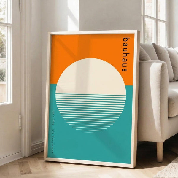 May include: Framed geometric poster leaning against white wall beside beige sofa in bright living room. Orange and teal color blocks with large cream circle containing white horizontal lines, resembling stylized sun over water. Black "bauhaus" text vertically aligned on right side. Light wood frame, approximately 2-3 feet tall, positioned on hardwood floor near large windows with natural lighting.