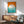 May include: Mid-century modern room setting with teal upholstered armchair, wooden sideboard, and white table lamp. Framed poster on white wall features orange and turquoise geometric design with white circle, horizontal lines, and "bauhaus" text. Natural sunlight creates shadows across the scene, highlighting the vintage aesthetic and clean lines of the furniture.