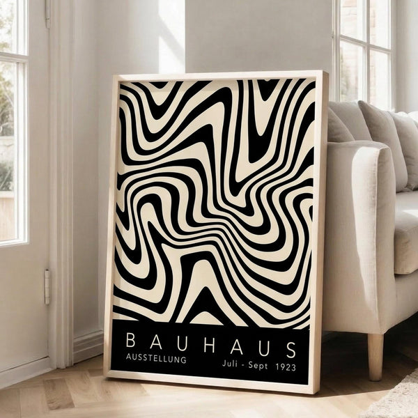 May include: Black and white abstract swirling pattern poster with curved flowing lines creating an optical illusion effect, displayed in light wood frame leaning against white wall next to beige upholstered sofa in bright living room with large windows and natural lighting on hardwood floors.