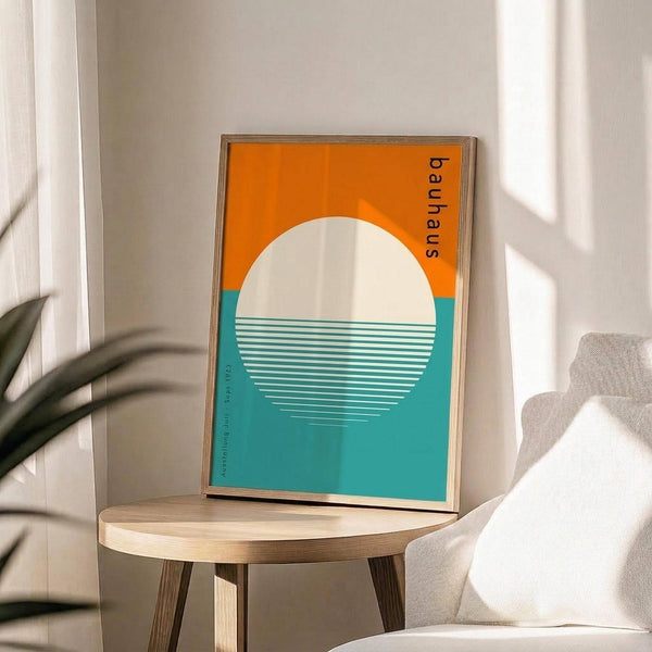 May include: Framed Bauhaus-style poster with geometric design featuring large white circle overlapping orange and teal sections with horizontal white stripes, displayed on round wooden side table next to beige upholstered chair, with plant leaves visible and natural window light creating shadows on white wall