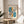 May include: Framed Bauhaus-style poster with geometric shapes in teal, orange, black and white displayed on wooden round stool next to beige upholstered chair. Abstract design features oval forms, rectangles, circles and linear patterns. Natural lighting creates soft shadows on white wall with plant leaves visible in corner of minimalist room setting.