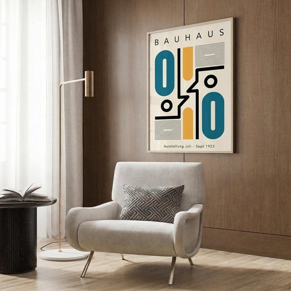 May include: Framed Bauhaus exhibition poster with geometric shapes in teal blue, orange, and black on cream background, displayed on wood paneled wall above beige upholstered armchair with geometric patterned pillow, next to brass floor lamp and black side table with open book