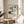 May include: Framed geometric art print with green and black abstract shapes including concentric circles, curved lines, and an eye motif on cream background, displayed on wooden side table next to beige upholstered chair with plant shadow cast on white wall behind