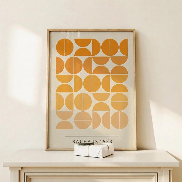 May include: Framed geometric art print with orange and yellow circular and semi-circular shapes arranged in a grid pattern, displayed on a light wood console table against a cream wall. The poster features "BAUHAUS 1923" text at bottom and gradual color transition from deep orange at top to pale yellow at bottom. Small wrapped gift box sits beside the natural wood frame.