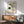 May include: Framed geometric art print with green circles and black concentric line patterns on cream background, displayed above mid-century modern wooden credenza with white ceramic lamp. Green upholstered armchair with wooden frame visible in foreground, natural sunlight streaming across white wall creating shadows.