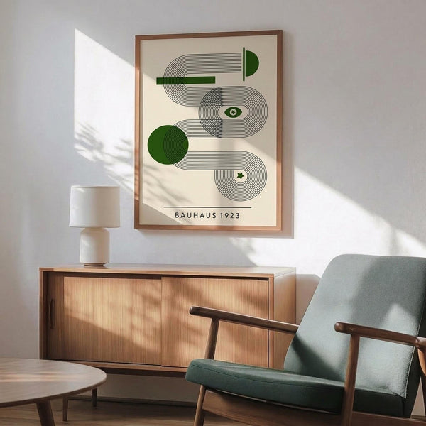 May include: Framed geometric art print with green circles and black concentric line patterns on cream background, displayed above mid-century modern wooden credenza with white ceramic lamp. Green upholstered armchair with wooden frame visible in foreground, natural sunlight streaming across white wall creating shadows.