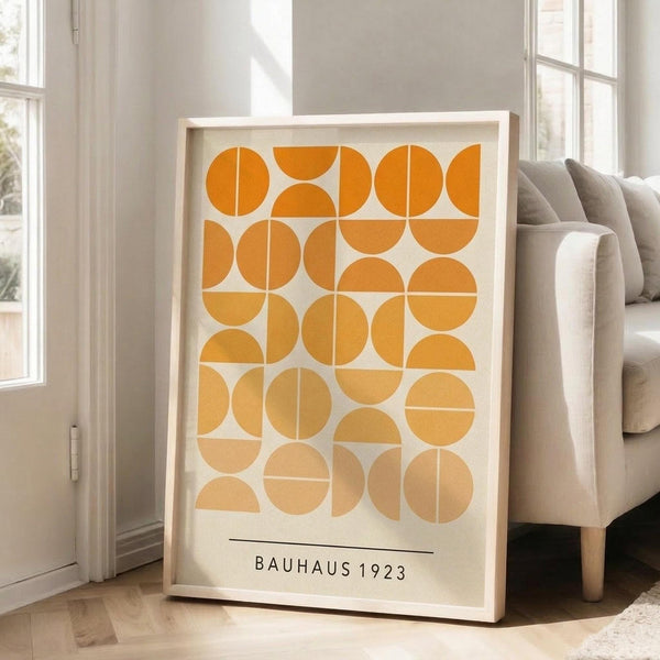 May include: Framed geometric art print with orange and yellow circles and semicircles arranged in a grid pattern on cream background, displayed on hardwood floor next to beige upholstered sofa in bright living room with white walls and large windows.