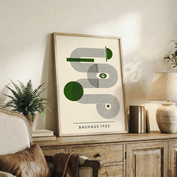 May include: Framed geometric abstract art print with green and black concentric circles and curved lines in Bauhaus style, displayed on wooden console table with books, ceramic vases, and fern plant in neutral living room setting