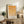 May include: Framed geometric art print with orange and yellow circular and semicircular shapes arranged in a grid pattern on white background, displayed on rustic wooden sideboard alongside fern plant, ceramic vase, and stacked books in warm, naturally-lit living room setting with beige upholstered chair.