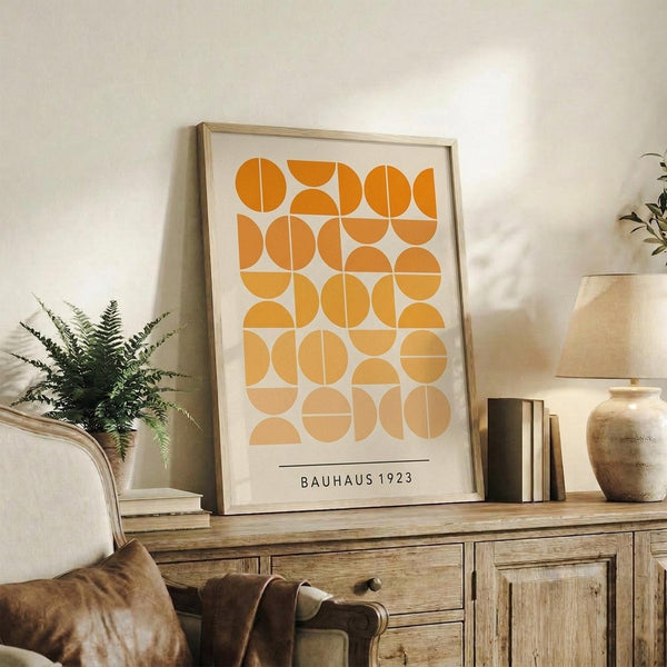May include: Framed geometric art print with orange and yellow circular and semicircular shapes arranged in a grid pattern on white background, displayed on rustic wooden sideboard alongside fern plant, ceramic vase, and stacked books in warm, naturally-lit living room setting with beige upholstered chair.