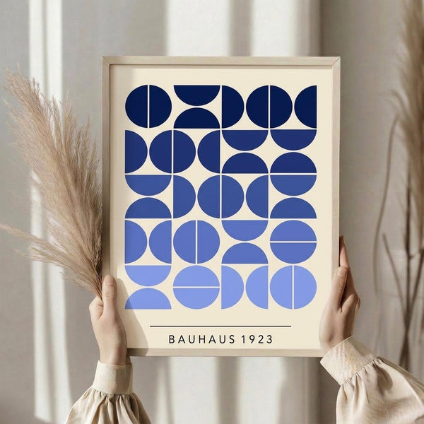 May include: Person holding framed geometric poster with blue circles and semicircles in gradient shades from navy to light blue on cream background, text reading "BAUHAUS 1923" below design. Light wood frame, neutral wall backdrop, dried pampas grass visible on left side.