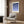 May include: Modern living room scene with framed geometric poster featuring blue circular shapes arranged in a grid pattern on cream background, displayed above beige upholstered armchair with patterned throw pillow. Room has wood paneled walls, brass floor lamp, and dark round side table with open book.