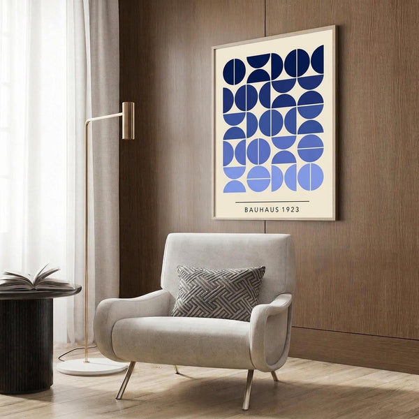 May include: Modern living room scene with framed geometric poster featuring blue circular shapes arranged in a grid pattern on cream background, displayed above beige upholstered armchair with patterned throw pillow. Room has wood paneled walls, brass floor lamp, and dark round side table with open book.
